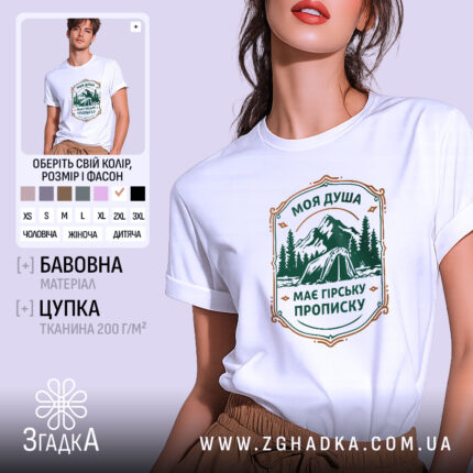 Футболка душа з гірською пропискою, біла, зображена на моделі з легким ракурсом, фон світло-фіолетовий, принт з горами та наметом в зелених тонах з текстом.
