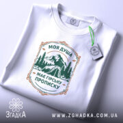 Футболка душа з гірською пропискою, біла, складена на столі, фон світлий, принт з горами та наметом в зелених тонах, прикріплена етикетка з зеленим шнурком.