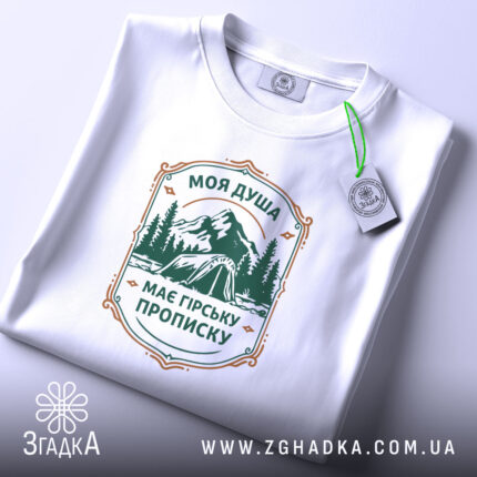 Футболка душа з гірською пропискою, біла, складена на столі, фон світлий, принт з горами та наметом в зелених тонах, прикріплена етикетка з зеленим шнурком.