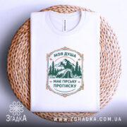 Футболка душа з гірською пропискою, біла, складена на плетеному кошику, фон світлий, принт з горами та наметом в зелених тонах, текст розташований у центрі.