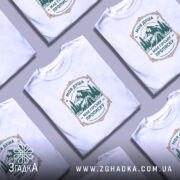 Футболка душа з гірською пропискою, біла, зображена на моделі з легким ракурсом, фон світло-фіолетовий, принт з горами та наметом в зелених тонах з текстом.