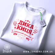 Футболка дика киця білого кольору, складена на поверхні, фон світло-сірий, принт червоного кольору з написом 'ДИКА КИЦЯ' та декоративними елементами, видима етикетка.