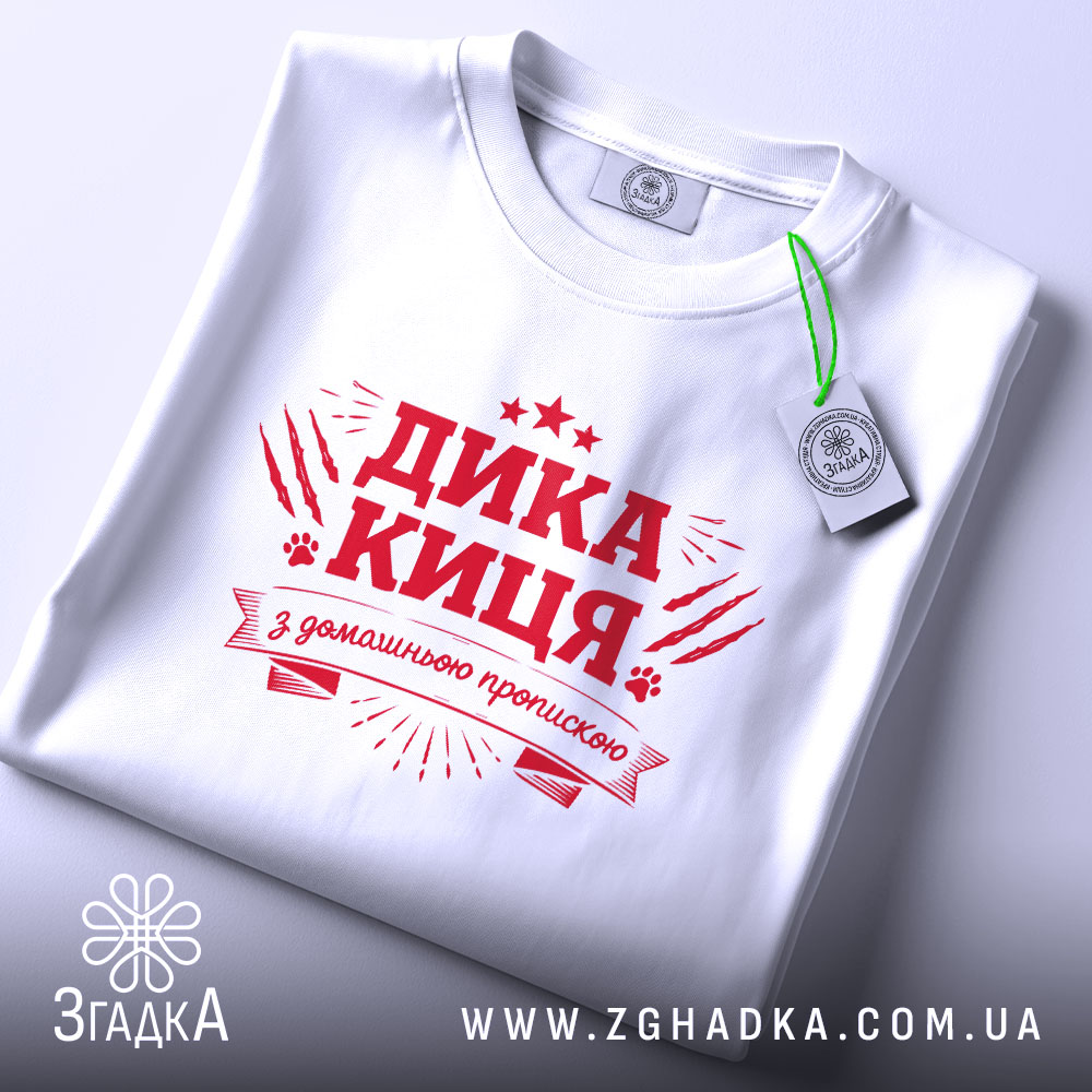 Футболка дика киця білого кольору, складена на поверхні, фон світло-сірий, принт червоного кольору з написом 'ДИКА КИЦЯ' та декоративними елементами, видима етикетка.