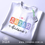 Футболка дихай вільно, біла, складена на столі, зображена зверху, фон світло-фіолетовий, принт з написом 'ДИХАЙ вільно' у кольорових квадратах, з сердечком над словом 'ДИХАЙ', на етикетці зелена стрічка.