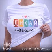 Футболка дихай вільно, біла, складена на столі, зображена зверху, фон світло-фіолетовий, принт з написом 'ДИХАЙ вільно' у кольорових квадратах, з сердечком над словом 'ДИХАЙ', на етикетці зелена стрічка.