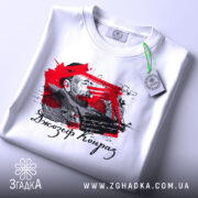 Футболка Джозеф Конрад, біла, складена на поверхні, фон світло-сірий, принт з червоними та чорними елементами, зображення чоловіка та текст, на етикетці зелена стрічка.