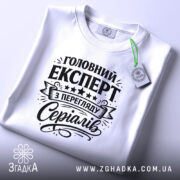 Футболка експерт з перегляду серіалів, біла, лежить на поверхні, фон світлий, принт чорного кольору з текстом 'Головний експерт з перегляду серіалів' та декоративними елементами, на комірці є етикетка.