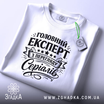 Футболка експерт з перегляду серіалів, біла, лежить на поверхні, фон світлий, принт чорного кольору з текстом 'Головний експерт з перегляду серіалів' та декоративними елементами, на комірці є етикетка.