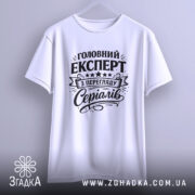 Футболка експерт з перегляду серіалів, біла, підвішена на вішалці, фон світло-сірий, принт чорного кольору з текстом 'Головний експерт з перегляду серіалів' та декоративними елементами.