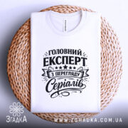 Футболка експерт з перегляду серіалів, біла, складена на плетеній основі, фон світлий, принт чорного кольору з текстом 'Головний експерт з перегляду серіалів' та декоративними елементами.