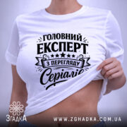 Футболка експерт з перегляду серіалів, біла, лежить на поверхні, фон світлий, принт чорного кольору з текстом 'Головний експерт з перегляду серіалів' та декоративними елементами, на комірці є етикетка.