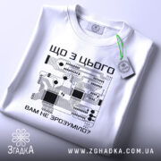 Футболка електрична схема, біла, зображена на плоскій поверхні, фон світло-сірий, принт з чорними елементами електронної схеми та текстом на українській мові, на комірці є етикетка.