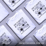 Футболка електрична схема, біла, зображена на моделі, що позує з легким усмішкою, фон світло-фіолетовий, принт з чорними елементами електронної схеми та текстом на українській мові.