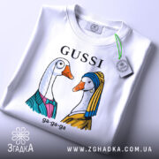 Футболка Gussi гуси білого кольору, складена на столі з нейтральним фоном. Принт з двома гусьми в одязі, одна з них у блакитному платті, інша в жовтому, з написом 'GUSSI' зверху. На футболці є етикетка.