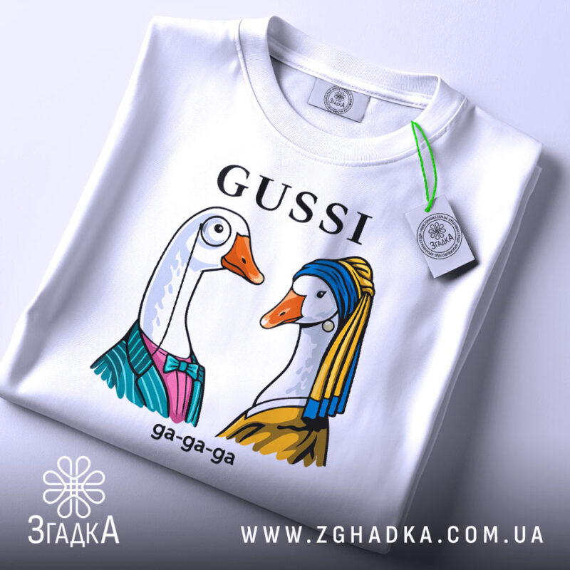 Футболка Gussi гуси білого кольору, складена на столі з нейтральним фоном. Принт з двома гусьми в одязі, одна з них у блакитному платті, інша в жовтому, з написом 'GUSSI' зверху. На футболці є етикетка.