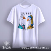 Футболка Gussi гуси білого кольору, підвішена на вішалці на світлому фоні. Принт з двома гусьми в одязі, одна з них у блакитному платті, інша в жовтому, з написом 'GUSSI' зверху.