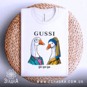 Футболка Gussi гуси білого кольору, складена на плетеній основі. Принт з двома гусьми в одязі, одна з них у блакитному платті, інша в жовтому, з написом 'GUSSI' зверху.