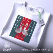 Футболка GUSSI з гусьми, біла, складена на поверхні, ракурс зверху, фон світло-сірий, принт з двома гусьми на червоному та зеленому фоні з написами GUSSI.