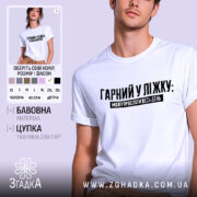 Футболка гарний у ліжку. Біла футболка на молодому чоловікові, зображена в анфас на світло-фіолетовому фоні. Принт чорного кольору з текстом "ГАРНІЙ У ЛІЖКУ: МОЖУ ПРОСПАТИ ВЕСЬ ДЕНЬ" розташований по центру. На футболці видно короткі рукави та округлий виріз.