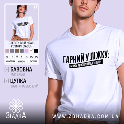 Футболка гарний у ліжку. Біла футболка на молодому чоловікові, зображена в анфас на світло-фіолетовому фоні. Принт чорного кольору з текстом "ГАРНІЙ У ЛІЖКУ: МОЖУ ПРОСПАТИ ВЕСЬ ДЕНЬ" розташований по центру. На футболці видно короткі рукави та округлий виріз.