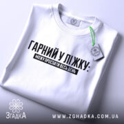 Футболка гарний у ліжку. Біла футболка, складена на поверхні, з видимим принтом чорного кольору. Текст "ГАРНІЙ У ЛІЖКУ: МОЖУ ПРОСПАТИ ВЕСЬ ДЕНЬ" розташований по центру. На футболці видно етикетку з зеленим шнурком.