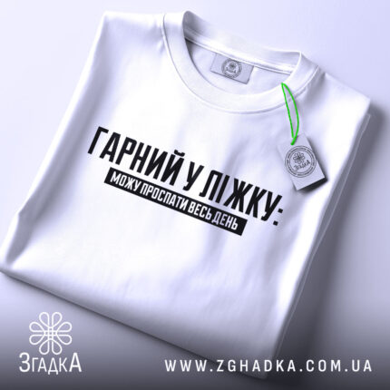 Футболка гарний у ліжку. Біла футболка, складена на поверхні, з видимим принтом чорного кольору. Текст "ГАРНІЙ У ЛІЖКУ: МОЖУ ПРОСПАТИ ВЕСЬ ДЕНЬ" розташований по центру. На футболці видно етикетку з зеленим шнурком.