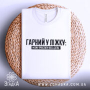 Футболка гарний у ліжку. Біла футболка, складена на плетеній основі, з видимим принтом чорного кольору. Текст "ГАРНІЙ У ЛІЖКУ: МОЖУ ПРОСПАТИ ВЕСЬ ДЕНЬ" розташований по центру. Фон світлий, що створює контраст з футболкою.