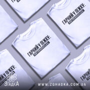 Футболка гарний у ліжку. Біла футболка на молодому чоловікові, зображена в анфас на світло-фіолетовому фоні. Принт чорного кольору з текстом "ГАРНІЙ У ЛІЖКУ: МОЖУ ПРОСПАТИ ВЕСЬ ДЕНЬ" розташований по центру. На футболці видно короткі рукави та округлий виріз.