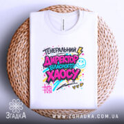 Футболка генеральний директор хаосу, біла, на столі, фон світло-сірий, принт кольоровий з написом 'ГЕНЕРАЛЬНИЙ ДИРЕКТОР ВЛАСНОГО ХАОСУ', розташована на плетеній основі, деталі принта яскраві.