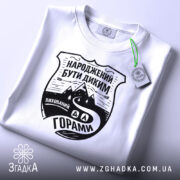 Футболка гірська свобода, біла, складена на поверхні, з ракурсом зверху, фон світло-сірий, принт чорного кольору з написом 'НАРОДЖЕНИЙ БУТИ ДИКИМ' та зображенням гір і дороги, з етикеткою зеленого кольору.