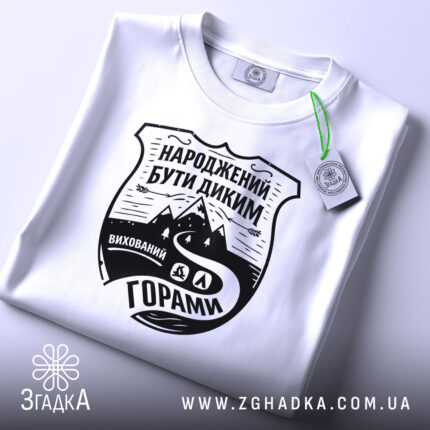 Футболка гірська свобода, біла, складена на поверхні, з ракурсом зверху, фон світло-сірий, принт чорного кольору з написом 'НАРОДЖЕНИЙ БУТИ ДИКИМ' та зображенням гір і дороги, з етикеткою зеленого кольору.