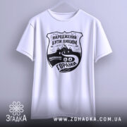 Футболка гірська свобода, біла, підвішена на вішалці, з ракурсом спереду, фон світло-сірий, принт чорного кольору з написом 'НАРОДЖЕНИЙ БУТИ ДИКИМ' та зображенням гір і дороги.