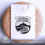 Футболка гірська свобода, біла, складена на плетеній основі, з ракурсом зверху, фон світло-сірий, принт чорного кольору з написом 'НАРОДЖЕНИЙ БУТИ ДИКИМ' та зображенням гір і дороги.