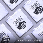 Футболка гірська свобода, біла, на моделі, з легким ракурсом спереду, фон світло-фіолетовий, принт чорного кольору з написом 'НАРОДЖЕНИЙ БУТИ ДИКИМ' та зображенням гір і дороги.