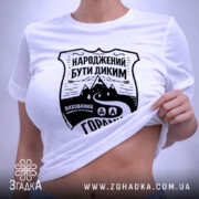 Футболка гірська свобода, біла, складена на поверхні, з ракурсом зверху, фон світло-сірий, принт чорного кольору з написом 'НАРОДЖЕНИЙ БУТИ ДИКИМ' та зображенням гір і дороги, з етикеткою зеленого кольору.