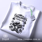 Футболка гори кличуть, біла, зображена на плоскості, ракурс зверху, фон світло-фіолетовий, принт чорного кольору з горами та туристичним спорядженням, з етикеткою зеленого кольору.