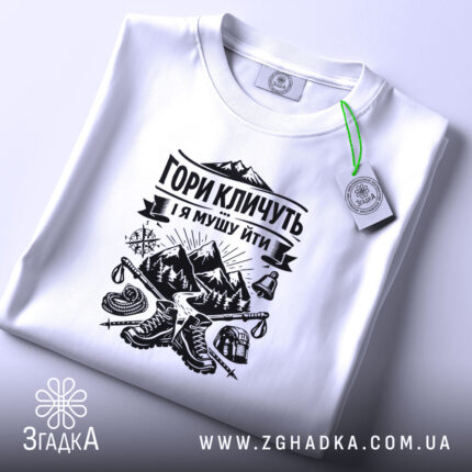 Футболка гори кличуть, біла, зображена на плоскості, ракурс зверху, фон світло-фіолетовий, принт чорного кольору з горами та туристичним спорядженням, з етикеткою зеленого кольору.