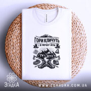 Футболка гори кличуть, біла, зображена на плоскості, ракурс зверху, фон світло-фіолетовий, принт чорного кольору з горами та туристичним спорядженням, на плетеній основі.