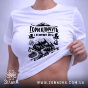 Футболка гори кличуть, біла, зображена на плоскості, ракурс зверху, фон світло-фіолетовий, принт чорного кольору з горами та туристичним спорядженням, з етикеткою зеленого кольору.