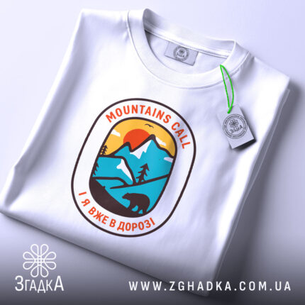 Футболка гори кличуть, біла футболка, складена на поверхні, принт у формі овалу з горами, сонцем та ведмедем, фон світло-сірий, на футболці є етикетка з зеленим шнурком.