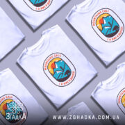 Футболка гори кличуть, біла футболка на моделі, з принтом у формі овалу, на якому зображені гори, сонце та ведмідь, фон світло-фіолетовий, деталі принта яскраві.