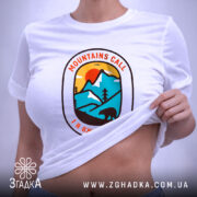 Футболка гори кличуть, біла футболка, складена на поверхні, принт у формі овалу з горами, сонцем та ведмедем, фон світло-сірий, на футболці є етикетка з зеленим шнурком.