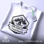 Футболка гори кличуть, біла, на плоскій поверхні, з етикеткою, фон світло-сірий, принт з горами, вогнищем і написом 'Гори кличуть! Я відповідаю.'