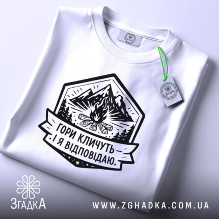 Футболка гори кличуть, біла, на плоскій поверхні, з етикеткою, фон світло-сірий, принт з горами, вогнищем і написом 'Гори кличуть! Я відповідаю.'
