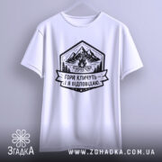 Футболка гори кличуть, біла, підвішена на вішалці, фон світло-сірий, принт з горами, вогнищем і написом 'Гори кличуть! Я відповідаю.'