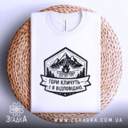 Футболка гори кличуть, біла, на плоскій поверхні, на плетеній основі, фон світло-сірий, принт з горами, вогнищем і написом 'Гори кличуть! Я відповідаю.'