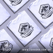 Футболка гори кличуть, біла, на моделі, з легким макіяжем, фон світло-фіолетовий, принт з горами, вогнищем і написом 'Гори кличуть! Я відповідаю.'