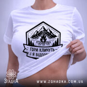 Футболка гори кличуть, біла, на плоскій поверхні, з етикеткою, фон світло-сірий, принт з горами, вогнищем і написом 'Гори кличуть! Я відповідаю.'