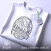 Футболка гори лікують. Біла футболка, складена на поверхні, фон світло-сірий, принт з горами та текстом чорного кольору, з етикеткою зеленого кольору.