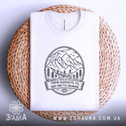 Футболка гори лікують. Біла футболка, складена на плетеній основі, фон світло-сірий, принт з горами та текстом чорного кольору.